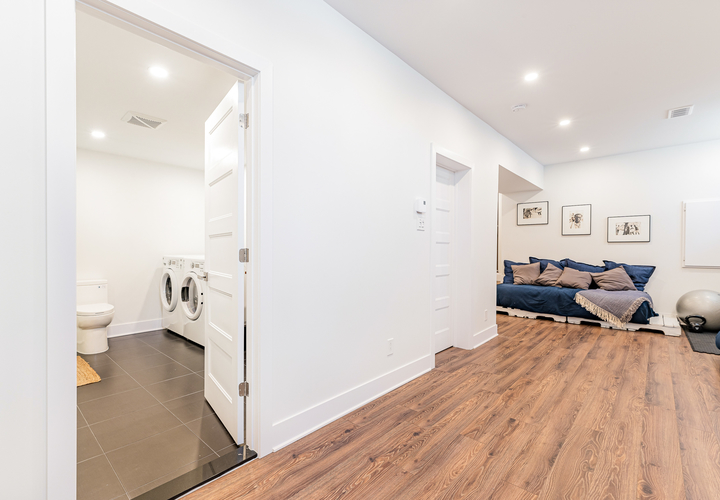A hallway with white walls and wooden floor leads to a bathroom with gray ceramic tiles, a washer, and a dryer