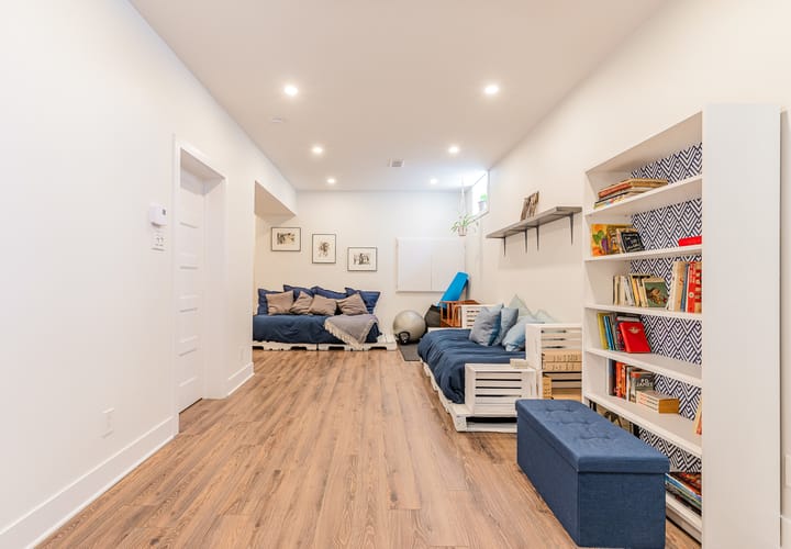 A bright basement with wooden floors, white walls, a bookcase, and two blue couches