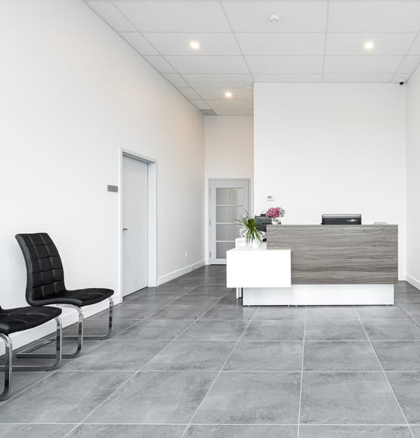 An office reception with black leather chairs, white walls, grey tiled floors and a two toned reception desk .