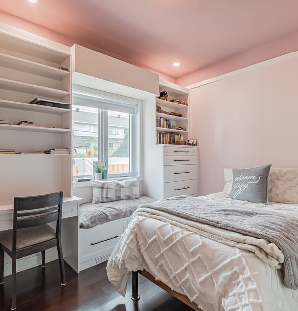 A light pink bedroom with white and grey bed, integrated desk and bookshelf with a window seat