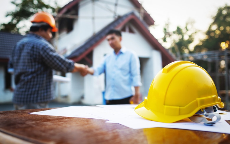 entrepreneur et client qui se serrent la main derrière un casque de construction jaune devant une maison en construction