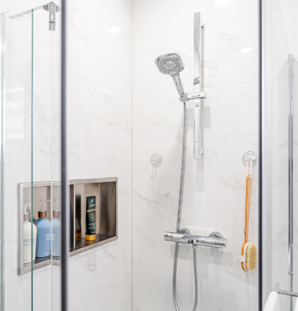 Renovated walk-in shower with faux white marble ceramic tiles, glass doors, silver hardware, and product niche