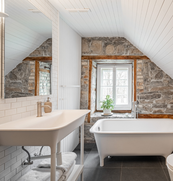 Salle de bain rustique rénovée avec bain autoportant sur pattes, vanité blanche et mur de pierre d'origine