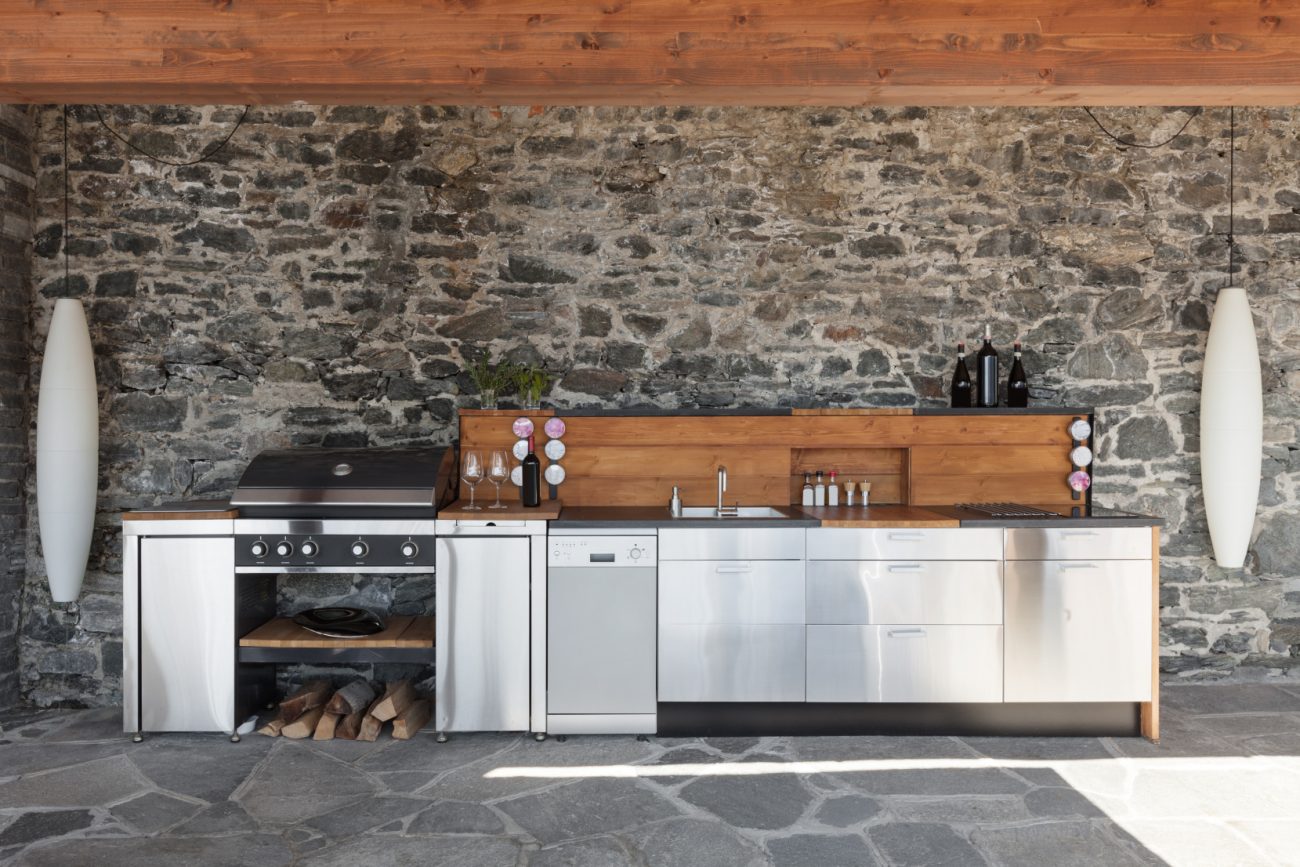 Outdoor kitchen with barbecue, sink, dishwasher and countertop, in front of a stone wall