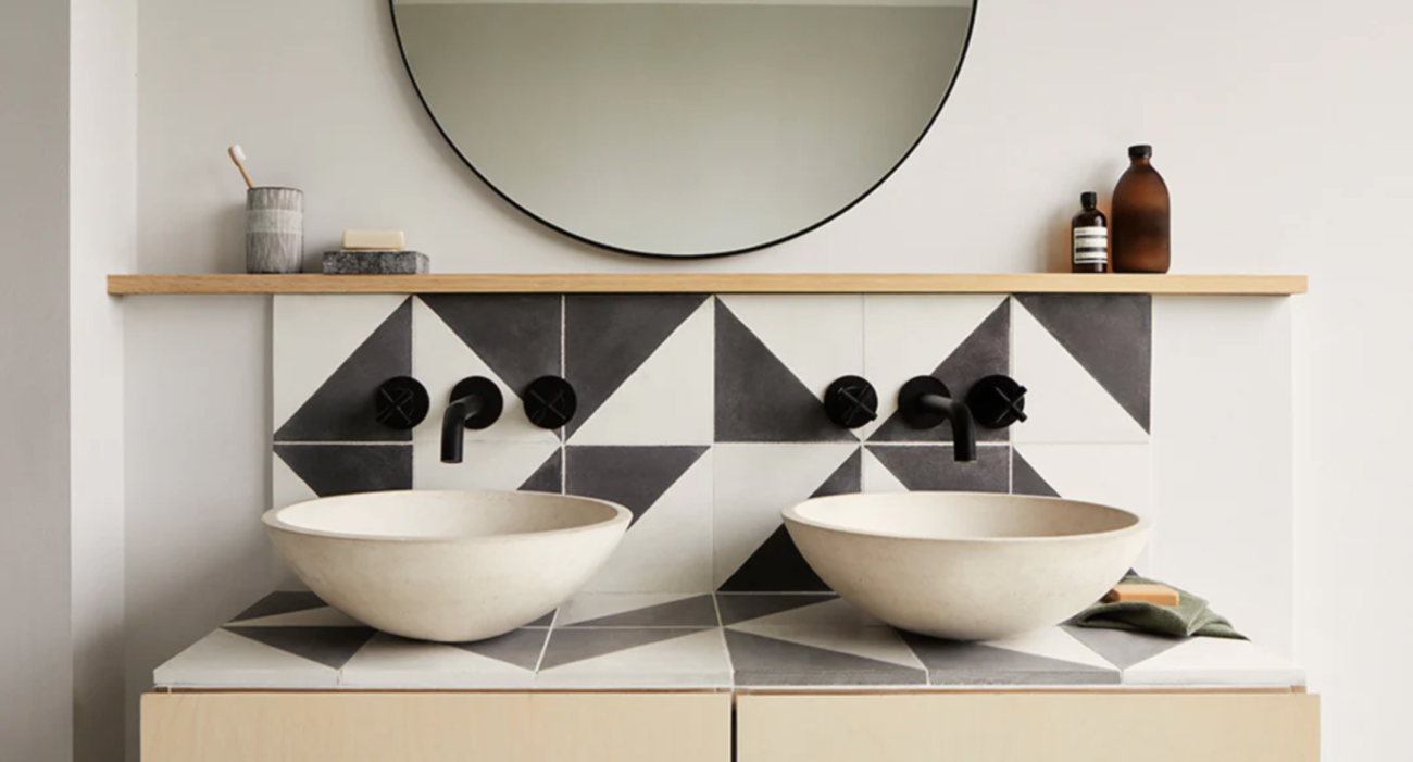 In a bathroom, two matte ceramic washbasins on a countertop decorated with black and white triangular tiles, round mirror, wooden shelf and black faucets