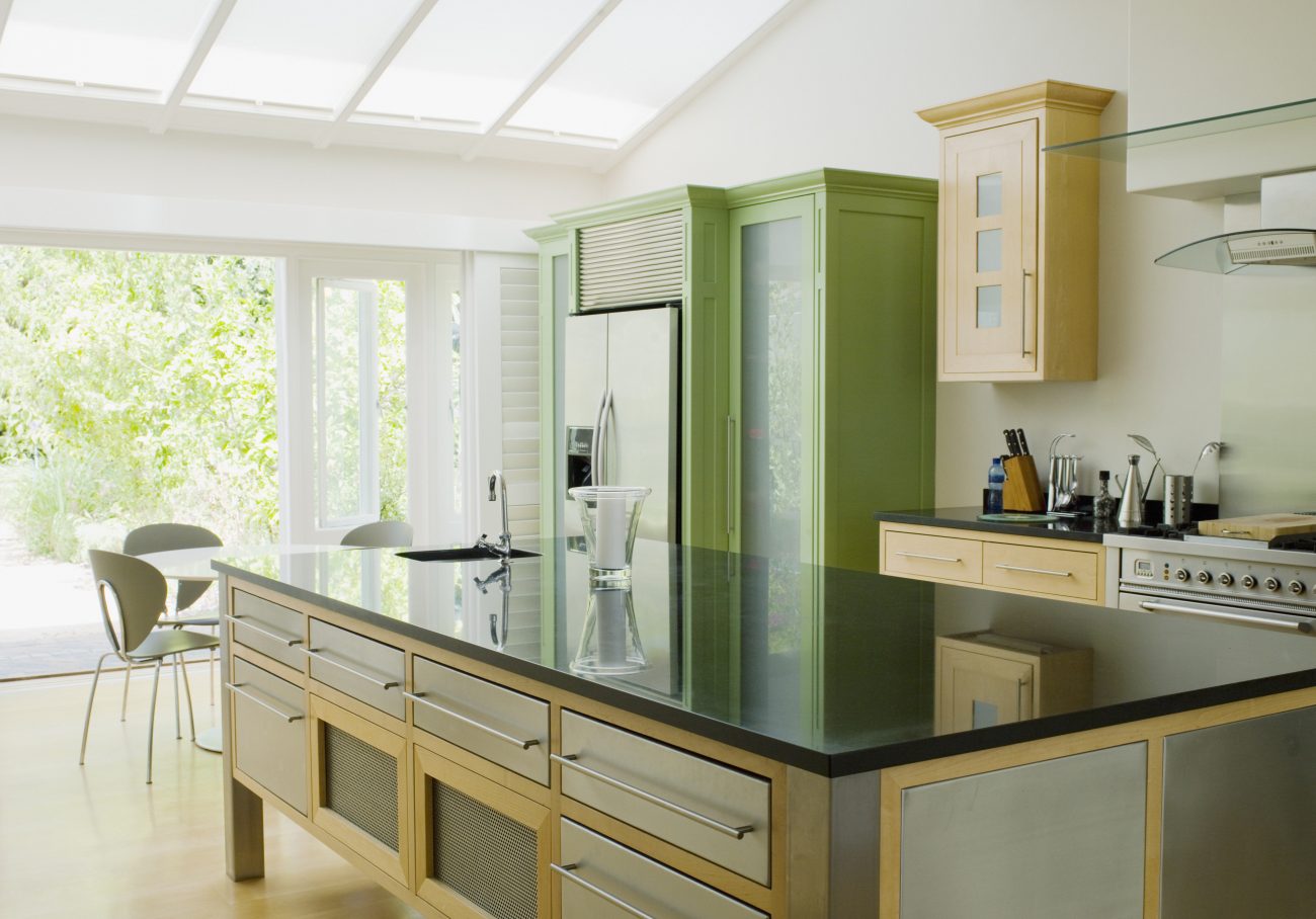 Cuisine lumineuse à portes-fenêtres et toit en verre, îlot à multiples tiroirs, armoire verte et rangement en bois pâle