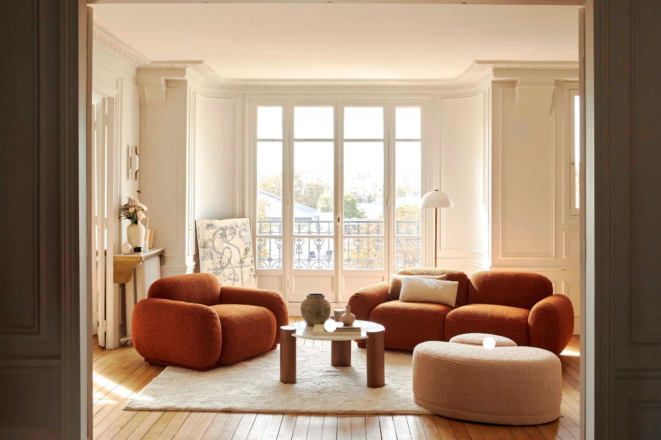 Sunny living room with large windowed doors, burnt orange bouclette wool sofa and armchair, beige ottoman and round coffee table