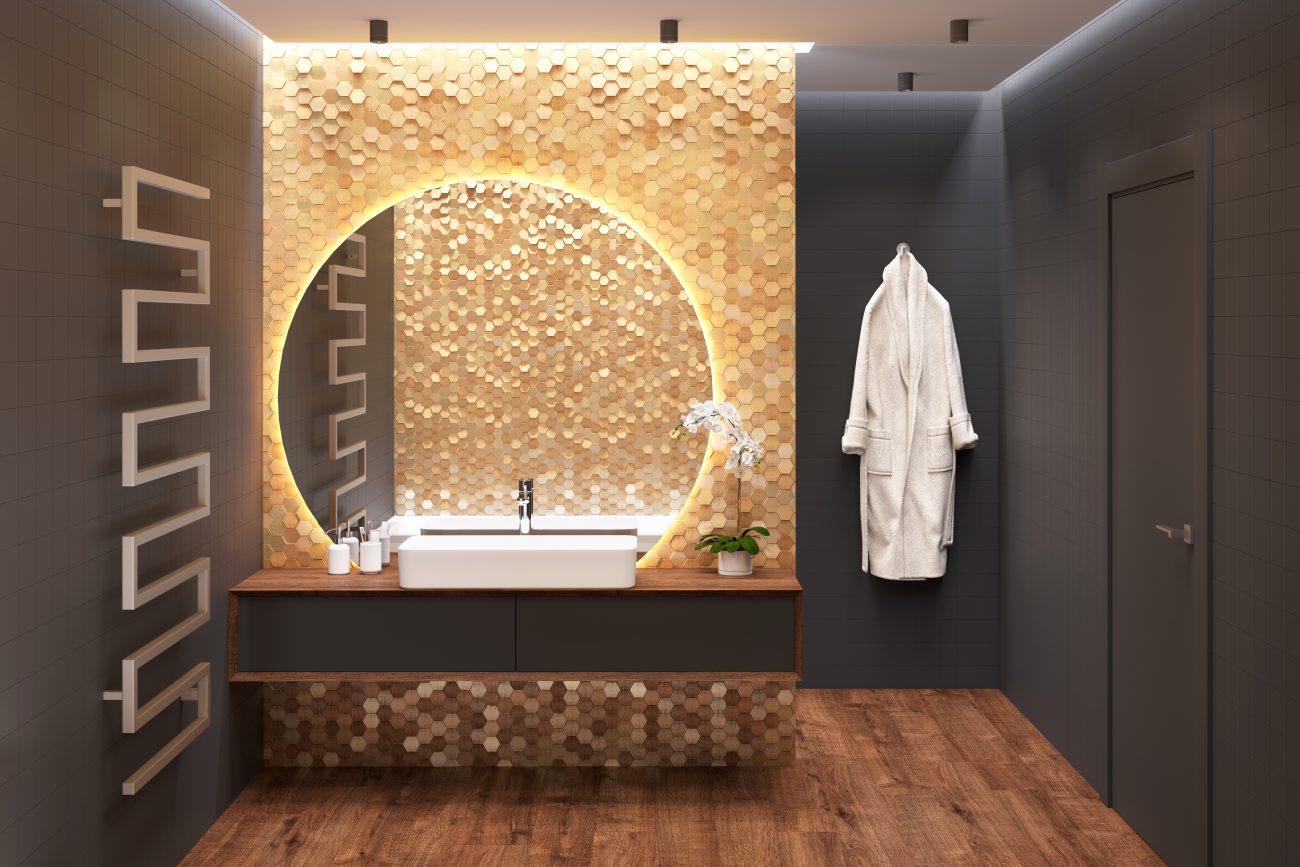 Salle de bain aux murs noirs, lavabo sur comptoir suspendu en bois, miroir rond rétroéclairé sur un mur en mosaïque dorée, porte-serviettes chauffant