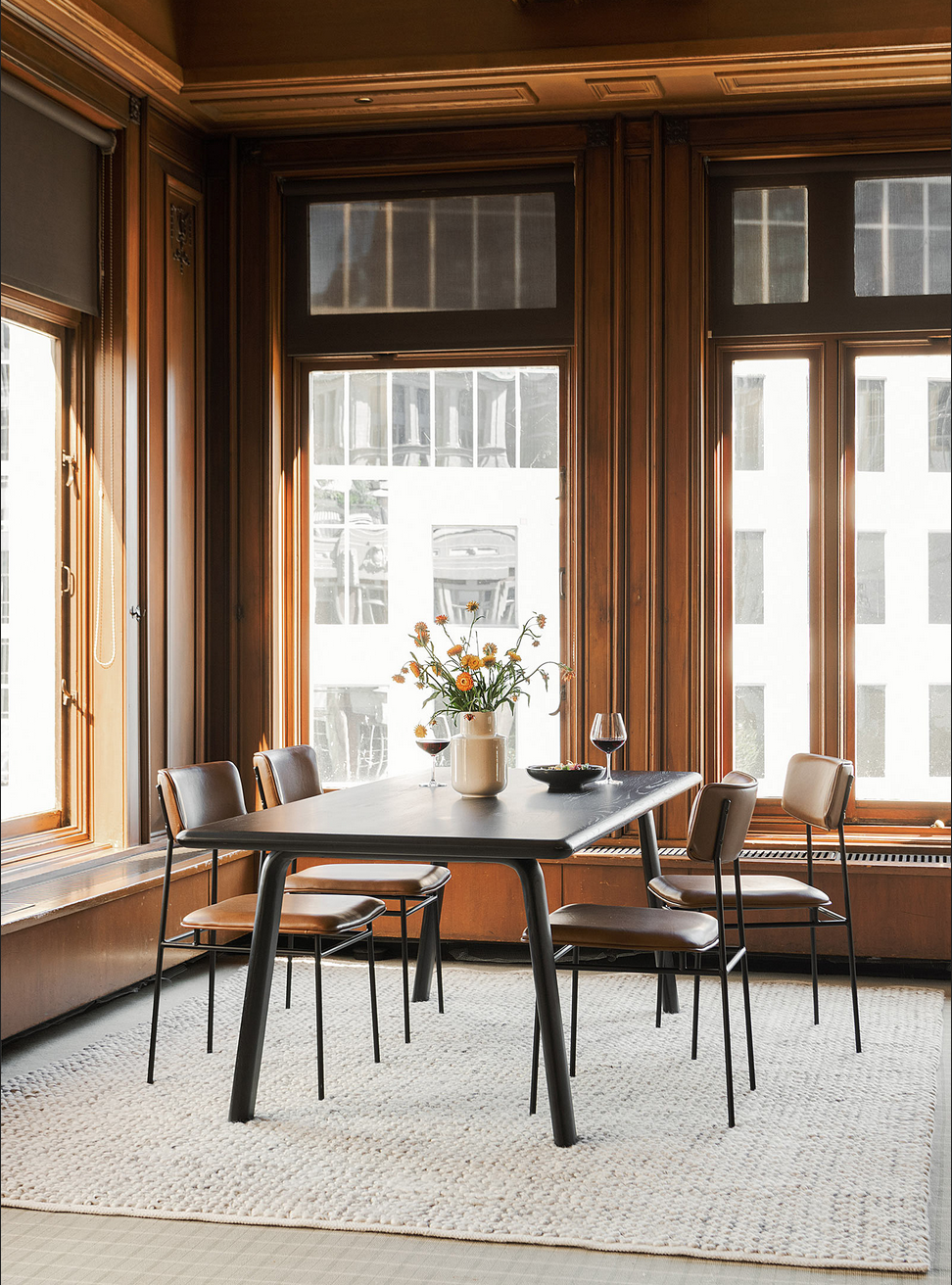 Salle à manger à plafond haut et grandes fenêtres en boiseries foncées, table rectangulaire en bois et chaises rétro en cuir brun, tapis texturé