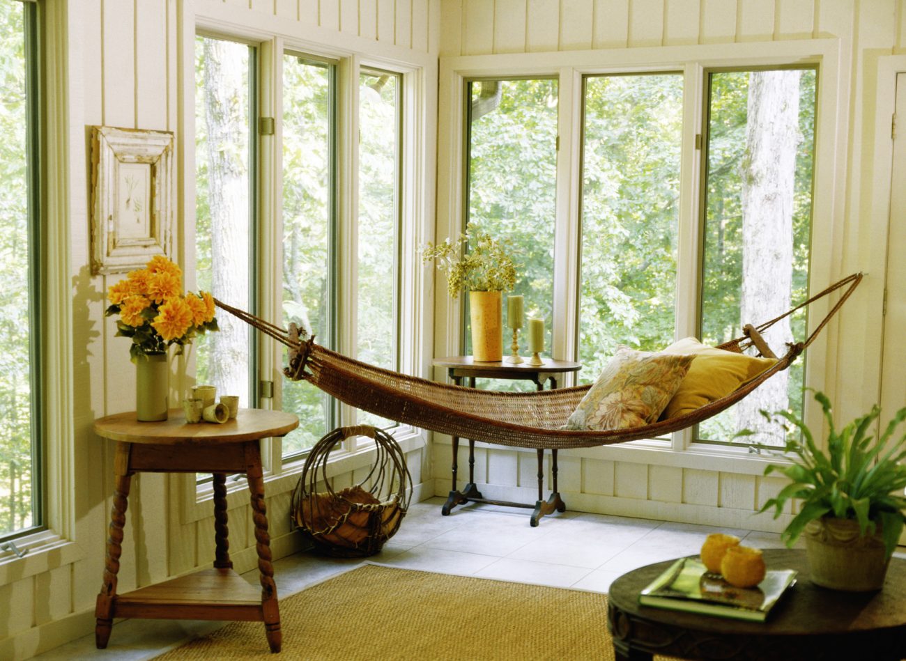 Sunroom surrounded by trees, wood-panelled walls, hammock, vintage wooden tables and flower vases