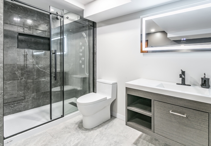 Modern bathroom in shades of gray and white with large shower including glass door and gray ceramic tiles, smart mirror, gray floating vanity, and dark faucets