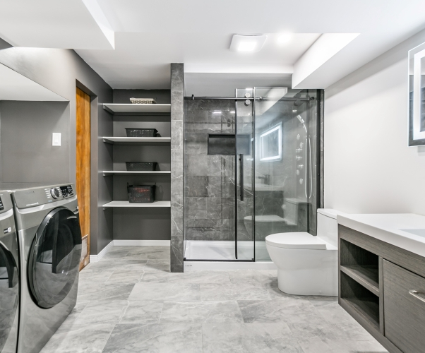 Transformation d’une salle d’eau en salle de bain complète de style contemporaine comprenant espace pour la lessive et rangements, le tout dans les teintes de gris et blanc.
