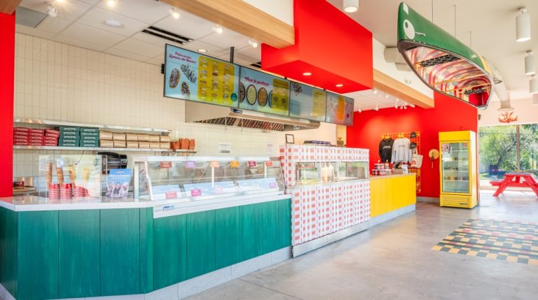 Renovated, traditionally inspired BeaverTails restaurant premises with bright colours (yellow, red, green), large windows, red benches and chairs, yellow, green and core-painted service counter, digital menu boards, concrete floor with a core-painted section, and a canoe suspended from the ceiling (yellow, red, green and core-painted).