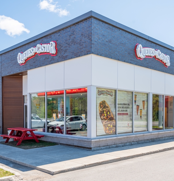 The exterior building of the BeaverTails franchise features a banner on the front and side wall, a red picnic table on the façade, large windows and walls painted white with a brick finish, and brown-walled entrance.