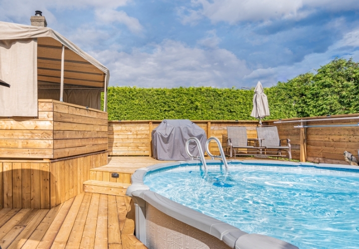 Terrasse sur trois paliers en bois traité de teinte naturelle avec auvent beige, chaises, balançoire et BBQ, entourant une piscine hors terre.