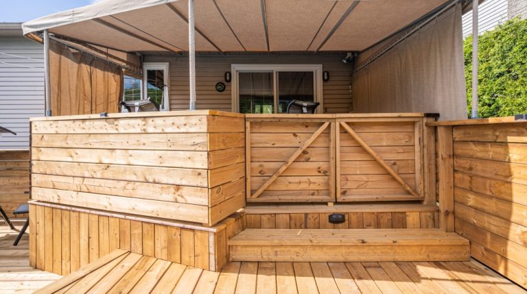 Porte-patio sur la façade arrière d’une maison avec terrasse sur trois paliers en bois traité teinte naturelle avec auvent en toile beige vissé dans le garde-corps.