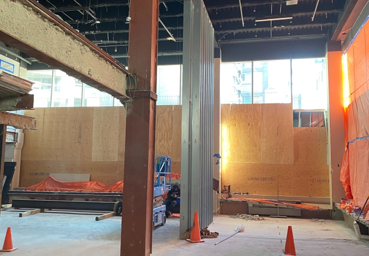 Refit work in a store, massive brown metal beams, dusty concrete floor with orange cones, boxes and various building materials, wall with large, plastered windows and another with large orange canvas, black metal ceiling.