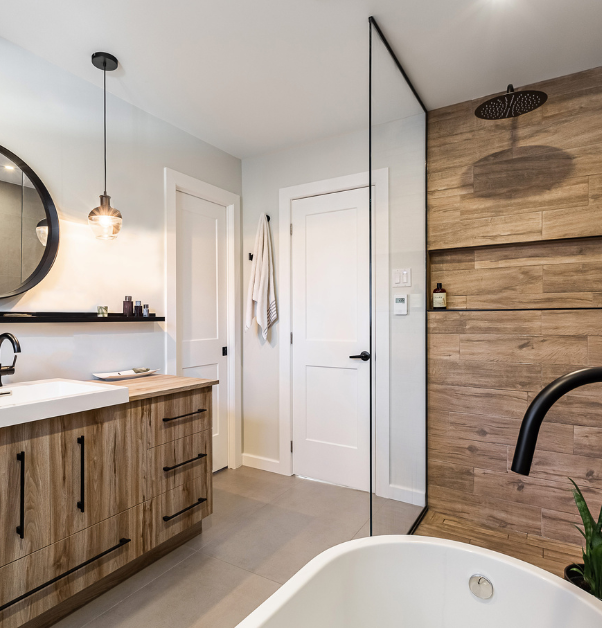 Renovated, contemporary-style bathroom painted white, white linen cupboard door and entrance door, glazed corner shower with a wall of the same laminate material as the natural wood imitation vanity, white washbasin with two cupboards and four drawers, black faucets, black oval mirror above the washbasin and a black shelf, black light fittings and sand-coloured ceramic floor.