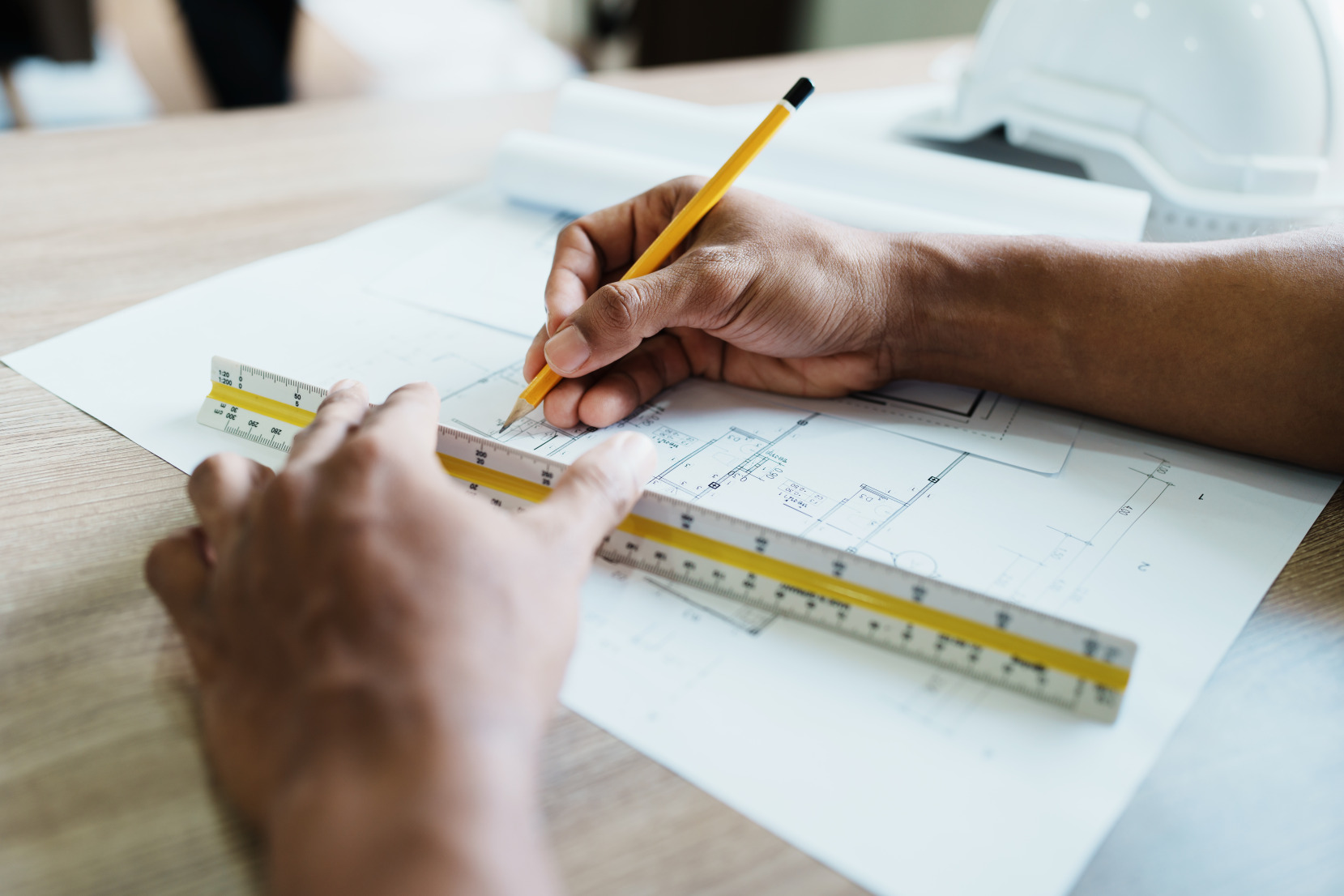 A close up of two brown hands using an architectural scale ruler and pencil to draw the floor plan of a home.