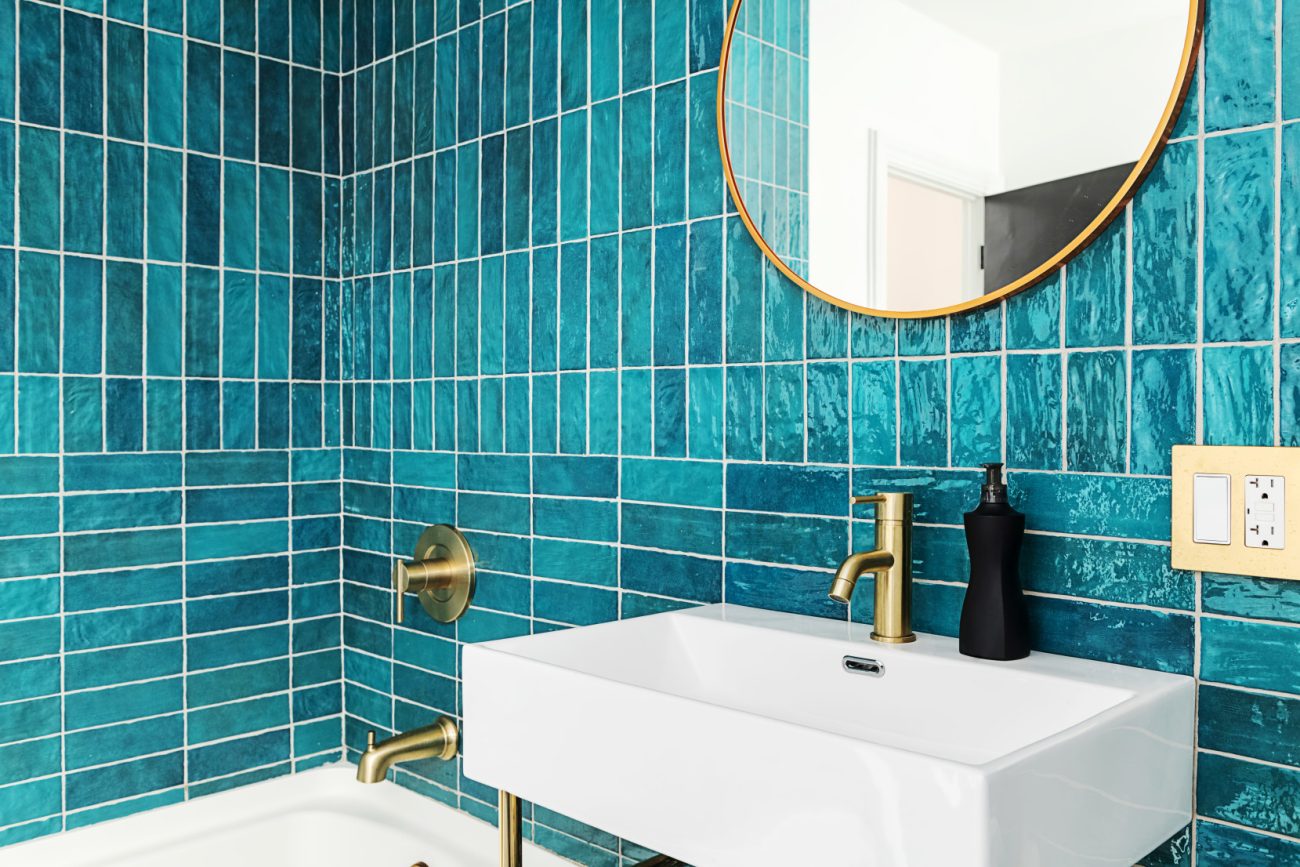 Bathroom with blue and green tiles with round gold mirror and faucets