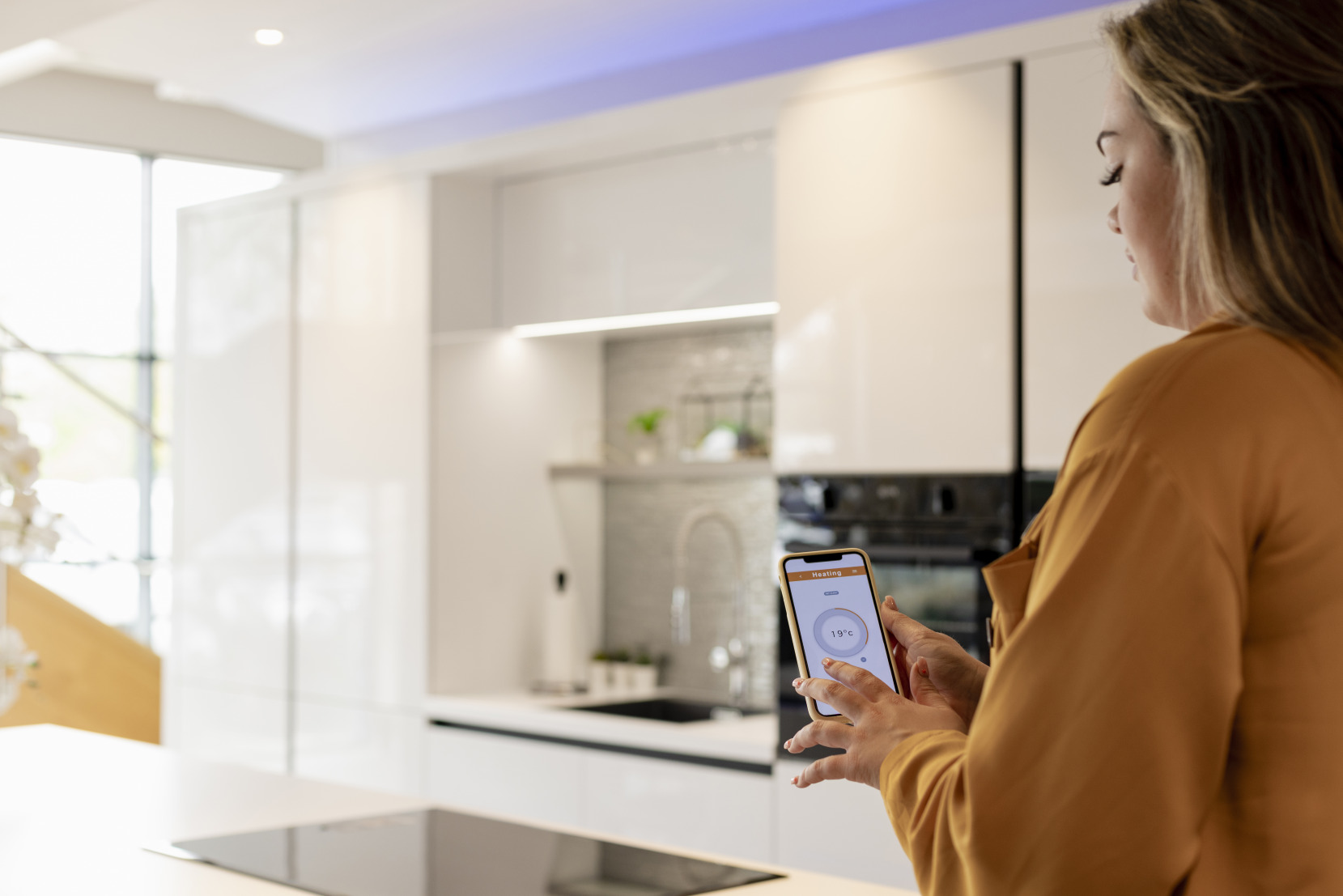 A homeowner stands in a contemporary white kitchen and controls home appliances using an app on their phone.