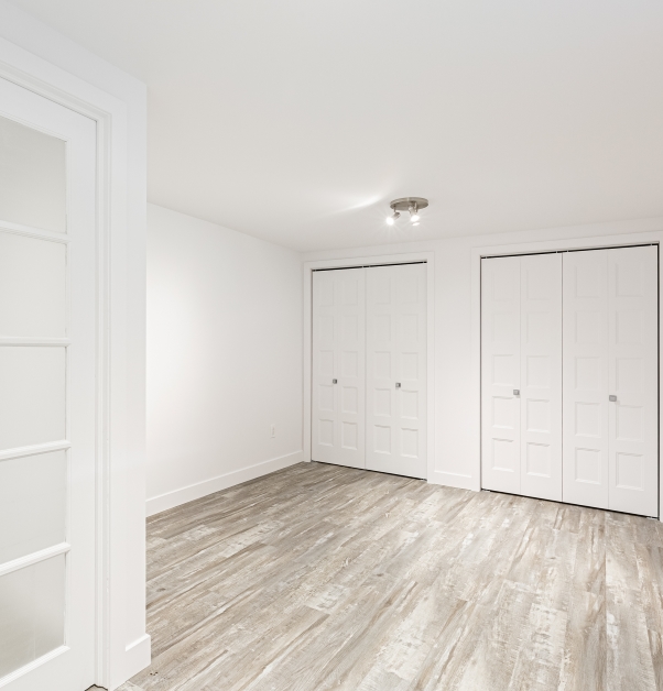 Grande chambre blanche avec plancher de vinyle imitation bois et deux garde-robes avec portes coulissantes blanches.