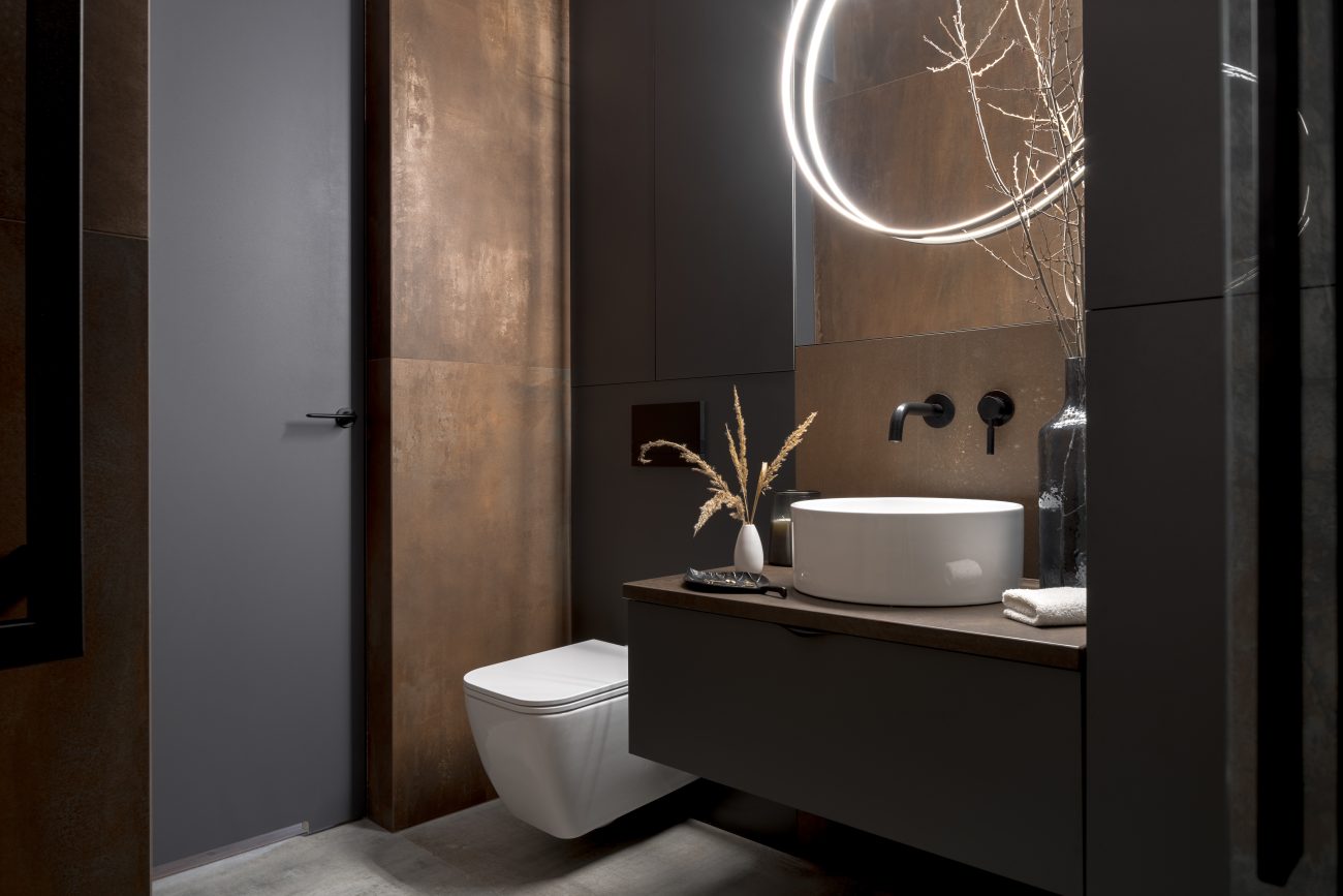 Modern bathroom with rust coloured tiles, black walls and illuminated round mirror