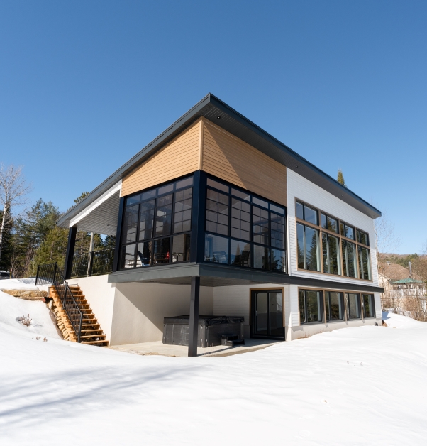 Maison moderne deux étages sur terrain en pente avec véranda vitrée noire, revêtement extérieur de bois d’ingénierie brun et blanc, rez-de-lac et toiture métallique en pente noire.