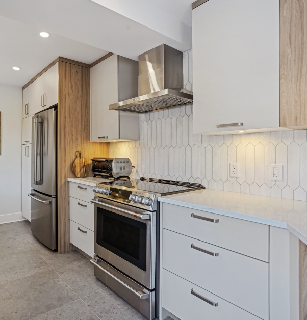 Modern renovated white kitchen with willow-gray and gold laminate cabinets, white ceramic backsplash, corner kitchen island with white stone countertop, stainless steel appliances and gray ceramic floor.