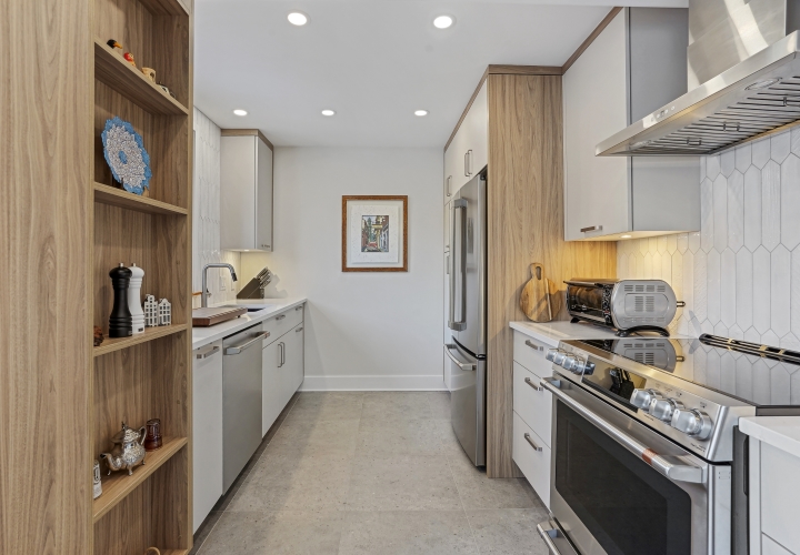 Renovated modern white kitchen with willow-gray and gold laminate cabinets, white ceramic backsplash, corner kitchen island with white stone countertop, black stools and gray ceramic floor.