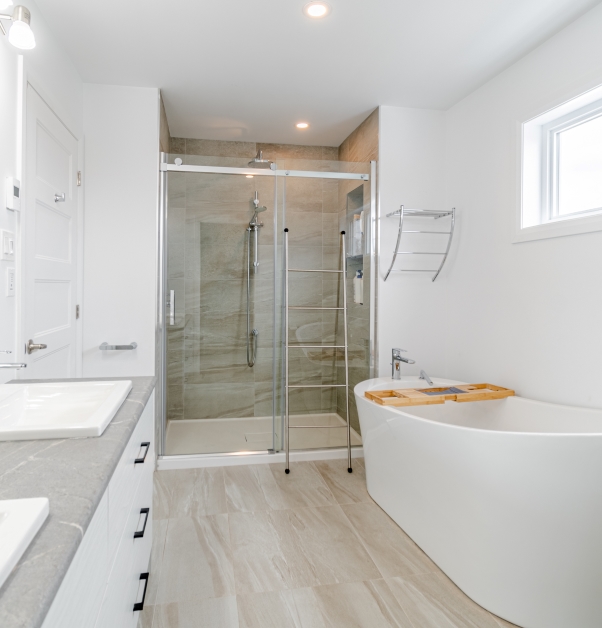 Classic white bathroom with white freestanding bathtub, glass shower with ceramic wall and floor, vanity with gray countertop and white cabinets, white sinks and beige ceramic floor.