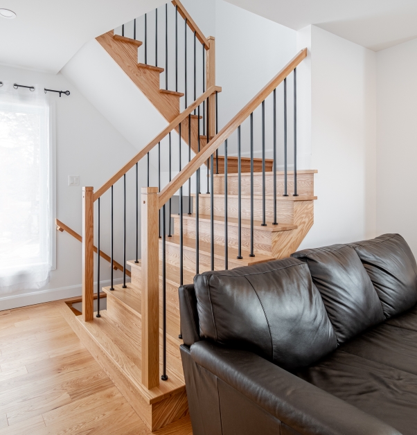 Cage d’escalier blanche avec marches et rampes en bois franc teinte naturelle, divan en cuir brun et grande fenestration blanche.